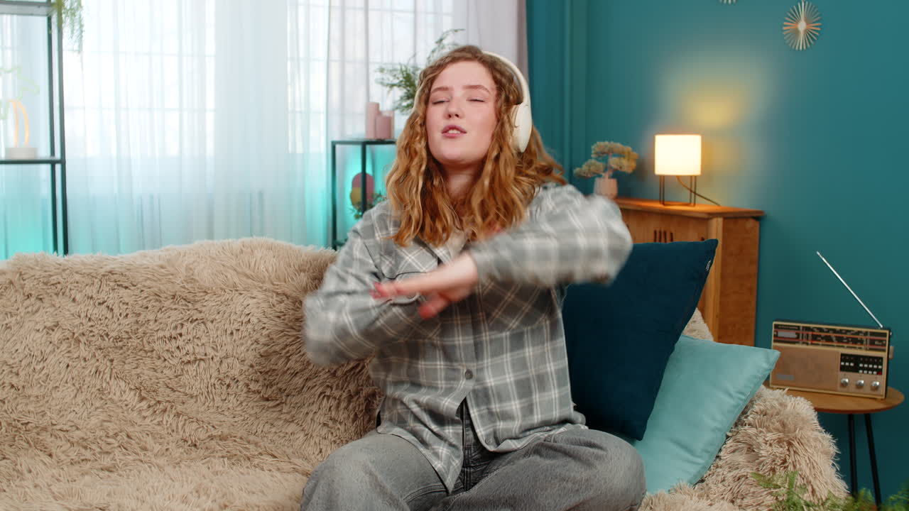 Woman sitting on sofa wearing headphones dancing with closed eyes enjoying favorite relaxing music