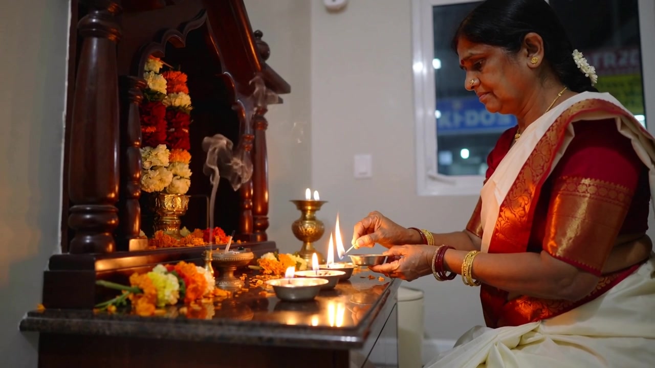 Una donna in sari color crema porta un piatto aarti acceso e accende delle diya sull'altare di casa mentre esegue la puja, con spazio per il testo.
