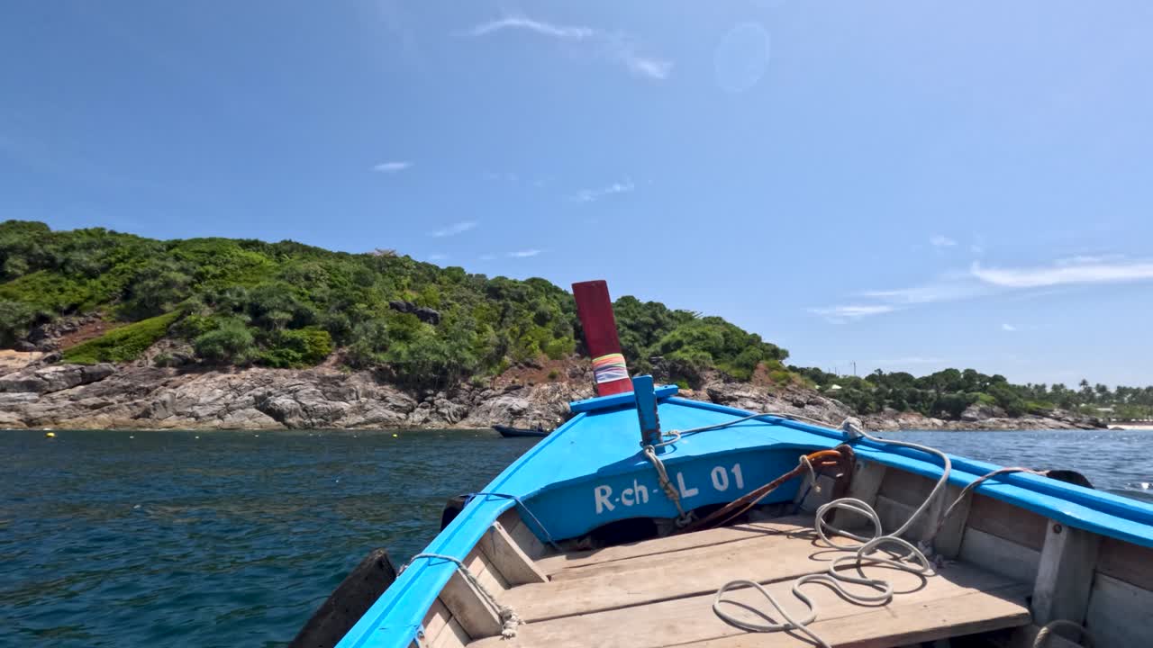 Wooden long-tail boat moves along rocky, green coastline under bright midday sun, steady forward perspective