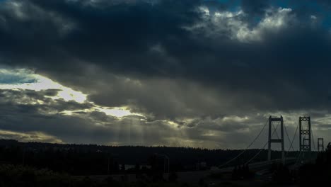 Time-lapse-of-the-sun-setting-over-the-Tacoma-Narrows-Bridge,-dramatic-sky,-sun-beams