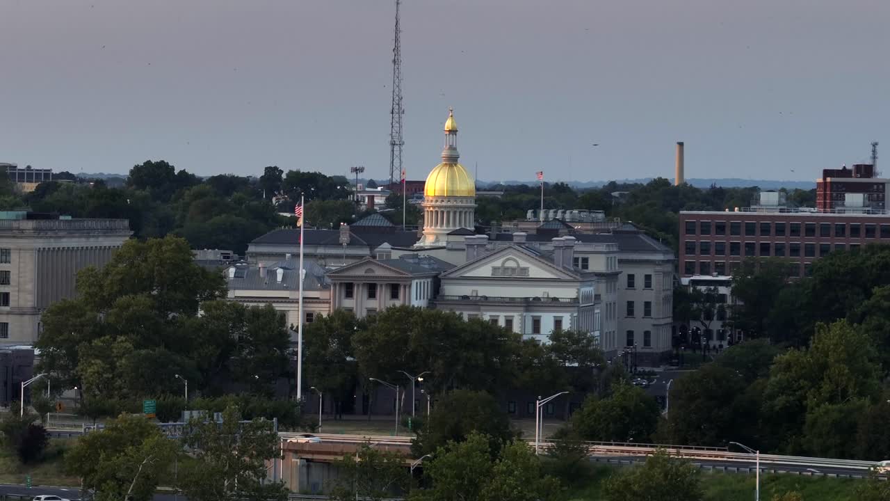 New York State capitol building with golden cupola in Trenton city. Cars on road along river. Waving American flag. Aerial rising wide shot