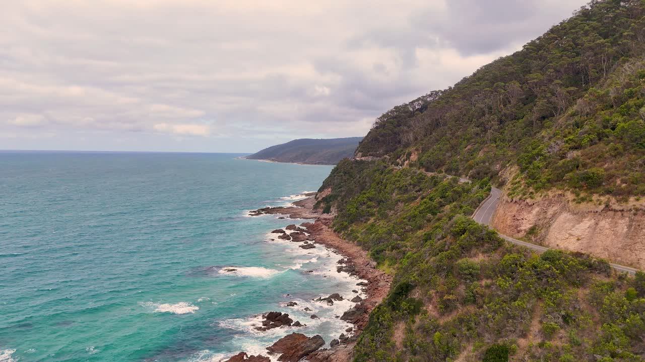 Drone footage captures the scenic coastal drive along Great Ocean Road, showcasing rugged cliffs and turquoise waters under overcast skies