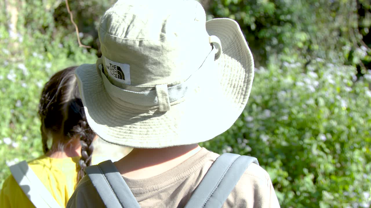 Duas crianças tailandesas pequenas usando mochilas e chapéus caminham por uma trilha ensolarada na floresta tropical