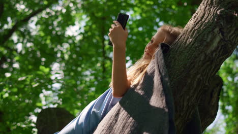 girl lying with phone on branch close up. smiling child watching smartphone.