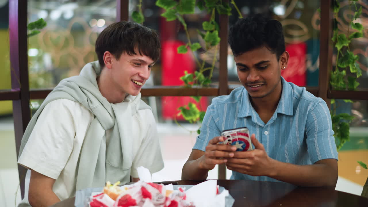 Colleagues laughing over video on phone with excitement on face while sharing tray of burger and chips, casual break scene with smiling peers, relaxed conversation and snack bonding moment