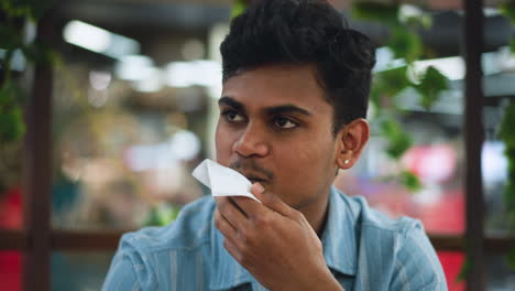 Adult wearing blue shirt taking bite of burger while smiling at his partner, closeup of happy colleagues sharing meal in casual setting, hands holding sandwich, background softly blurred