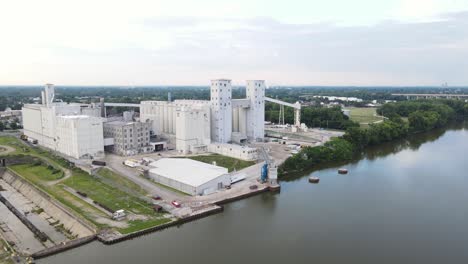 Luftaufnahme-Einer-Großen-Getreideaufzugsanlage-Entlang-Eines-Flusses-An-Einem-Leicht-Bewölkten-Sommertag-Mit-Ruhigem-Wasser,-In-Dem-Sich-Die-Bäume-Im-Wasser-Spiegeln