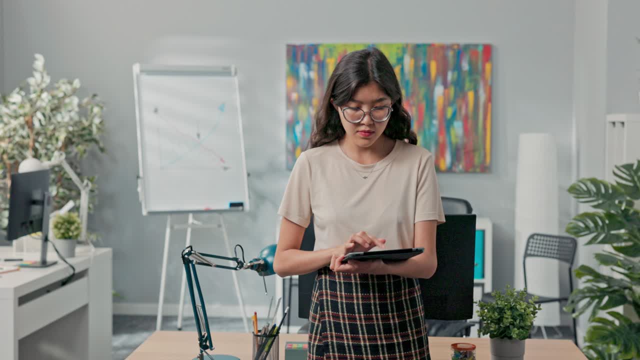안경을 쓴 아름다운 여성이 사무실 한가운데에 있는 책상 앞에 서서 타블에서 회사의 주문, 완료해야 할 주문의 수, 동료들에게 임무를 부여하고 있습니다.