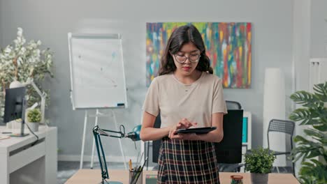 una hermosa mujer con gafas está de pie frente al escritorio en el medio de la oficina, comprobando en la tableta las órdenes de la compañía, el número de órdenes a completar, asignando tareas a los compañeros de trabajo