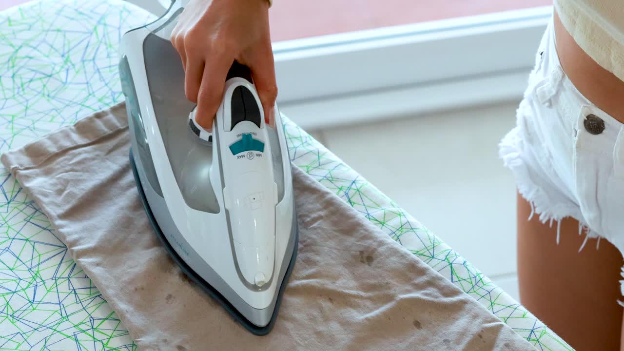 A woman uses a steam iron to press beige trousers on an ironing board in a sunlit, modern interior. Camera remains steady, natural daylight