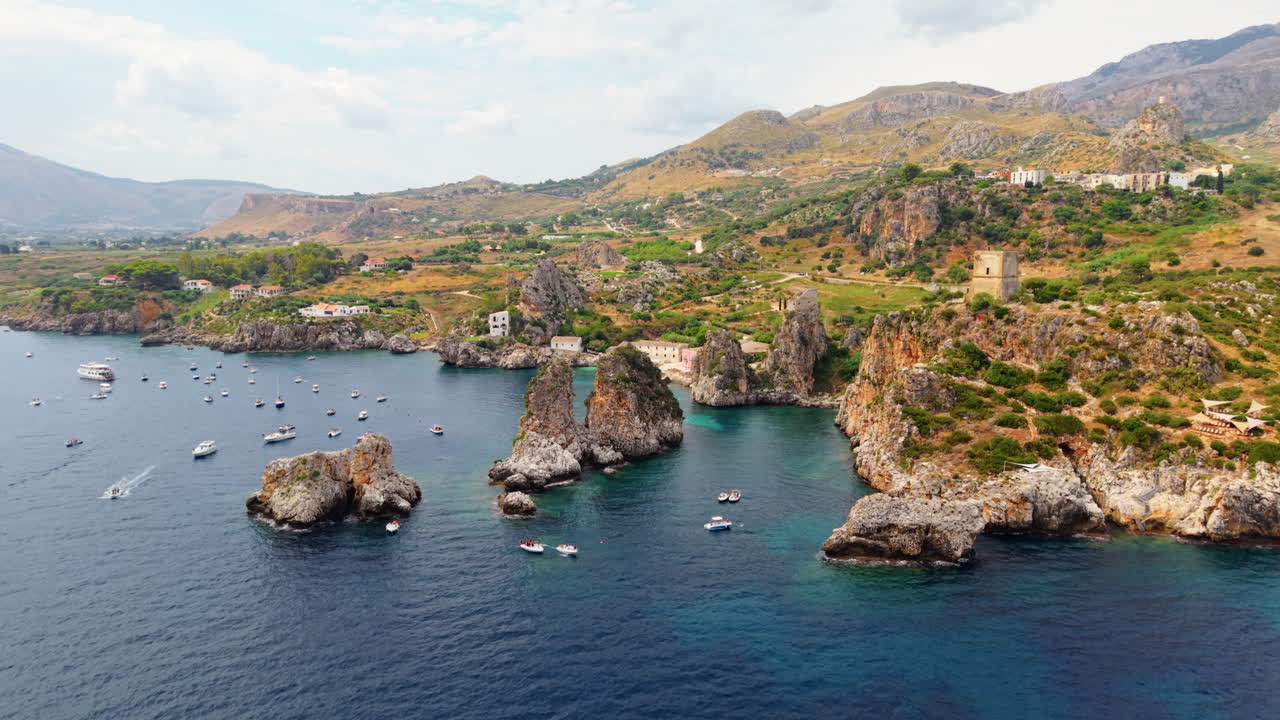 Drone ascends over the bay of Scopello, Sicily, revealing rocky cliffs, turquoise waters, and boats moving through the marina along the coast