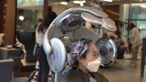 Young-Korean-woman-wearing-face-mask-and-eye-protection-sits-under-heating-lamps-inside-hair-salon,-static