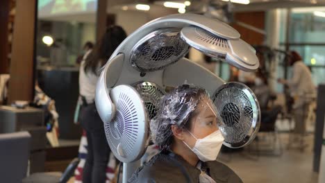 joven coreana con mascarilla y protección para los ojos se sienta bajo lámparas de calefacción dentro de la peluquería, estática