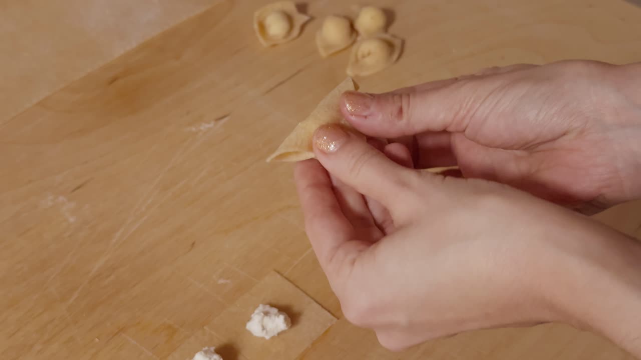 Primo piano di mani che piegano un quadrato ripieno di ricotta per formare cappelletti su una superficie di legno con illuminazione soffusa.