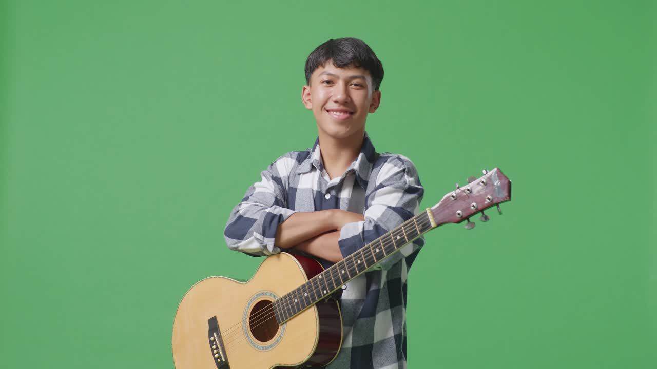 기타를 들고 있는 아시아 청소년이 팔을 고 초록색 스크린 배경에 미소 짓고 있습니다.