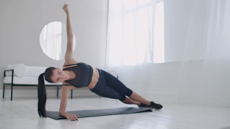 The-brunette-in-the-apartment-does-an-exercise-plank-while-standing-in-a-static-position-on-the-lateral-muscles-of-the-abs-and-moving-her-hand-up-and-down