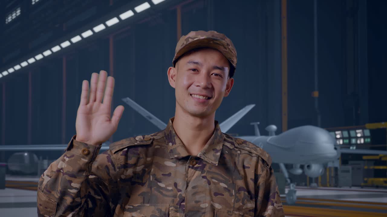 Primo piano di un soldato asiatico che sorride, saluta con la mano e dice addio, mentre si trova con un moderno drone militare in un hangar di manutenzione.