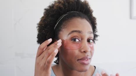 Happy-african-american-woman-applying-under-eye-mask-looking-in-bathroom-mirror,-slow-motion