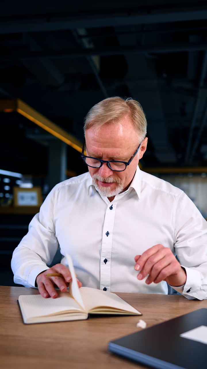 Adult male entrepreneur in white shirt sits at desk turning the pages of his notebook. Man says something opening his mouth wide and takes notes to his daybook. Vertical video.