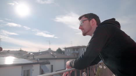 Young-attractive-caucasian-man-with-beard-and-glasses-on-balcony,-sunshine-pointing-at-happening-in-distance,-watching-people-below-him