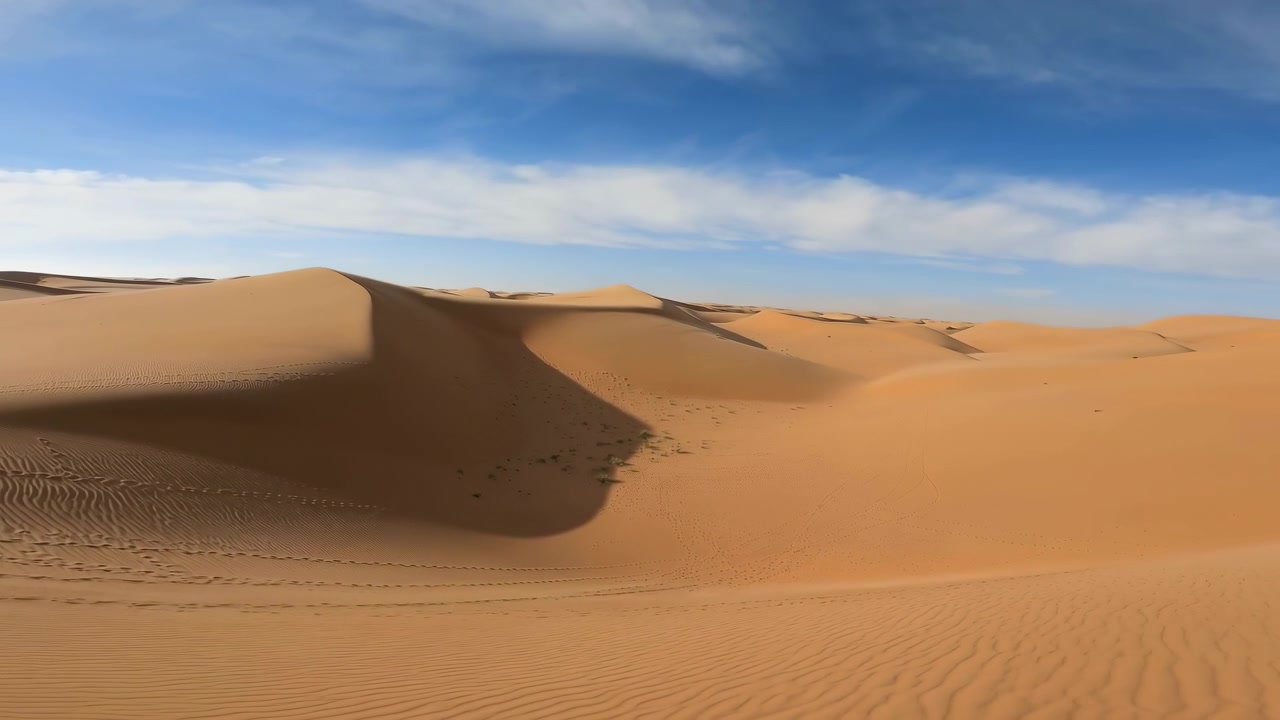 Ampio paesaggio desertico mostra dune dorate ondulate sotto un cielo azzurro brillante, con una profonda ombra centrale che aggiunge contrasto, texture e profondità attraverso il terreno arido, sereno ed espansivo, con deserto, duna