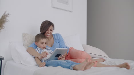 Mom-and-son-looking-at-the-tablet-screen-lying-on-a-white-bed.-Play-games-with-your-son-on-your-tablet-computer-and-watch-funny-videos.