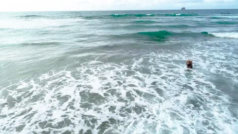 Ein-Mann-Und-Eine-Frau-In-Badeanzügen-Genießen-Heißes-Wasser-Am-Strand-Von-Neuseeland,-Wo-Die-Wellen-Auf-Das-Blaue-Wasser-Und-Den-Himmel-Krachen,-Die-Felsnadeln-Und-Der-Spaß-–-Ein-Dolly-Aus-Der-Luft