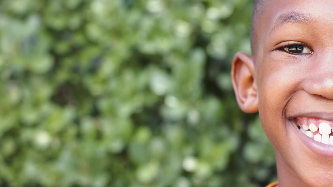 Bambino africano che sbircia da destra mentre la telecamera si sposta a sinistra, sorridendo contro una siepe verde, spazio per il testo