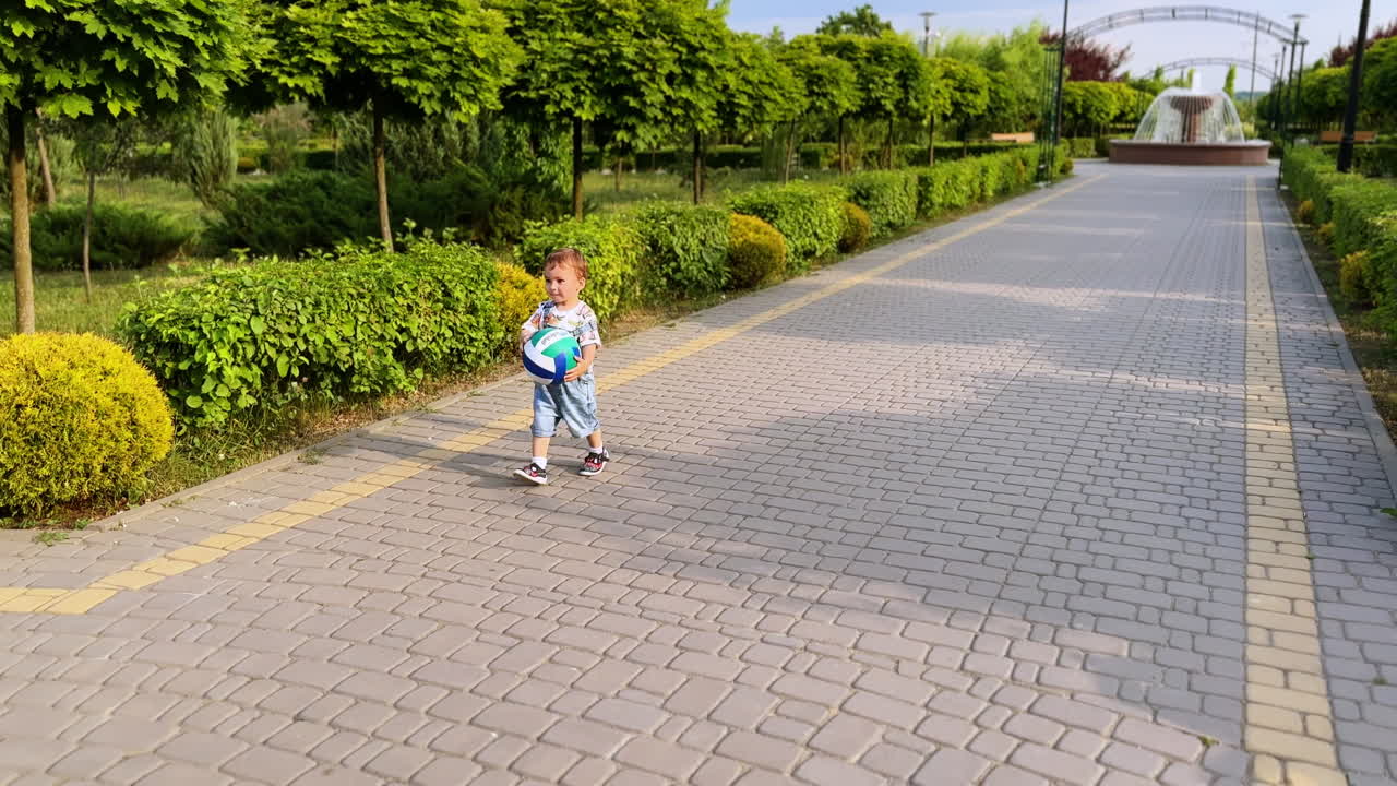 Adorable little child holding a ball runs by the alley in the park. Kid throws the ball and follows it.