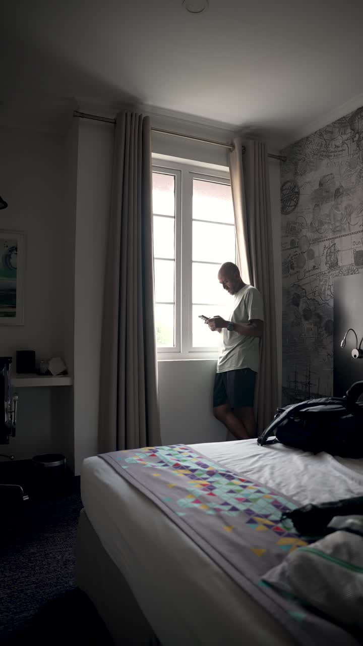 un turista está de pie junto a la ventana de su habitación de hotel, usando su teléfono inteligente y disfrutando de la vista