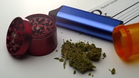 a small pile of ground marijuana lies on table surrounded by a grinder, vaporizer, open pill bottle and prescription note