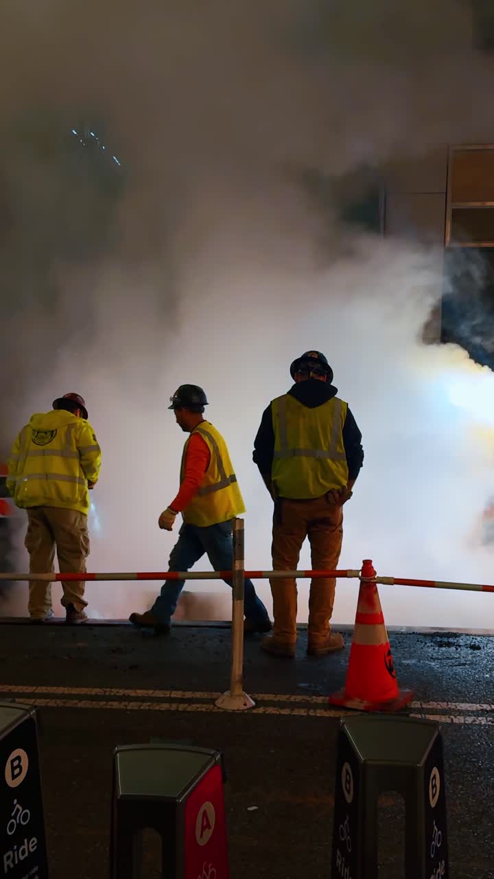 New York, USA, 25 April 2025: Utility workers standing by steaming manhole on NYC street night. Two construction workers in high visibility vests observing steam rising from a road manhole at night