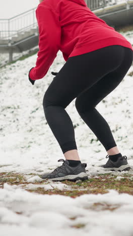 woman performing jump squats outdoors during winter workout on snowy ground wearing red hoodie and black leggings beside iron railing and staircase on snowy hill with bag in background