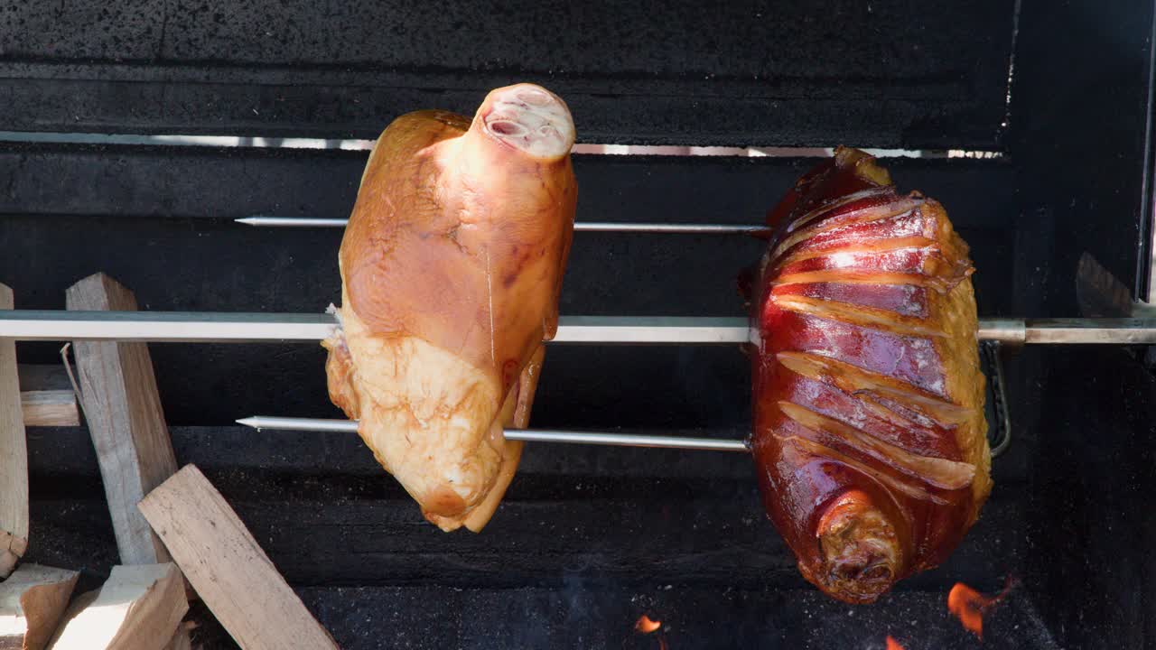 Two pork knuckles slowly rotate over open flames on a wood-fired spit grill outdoors