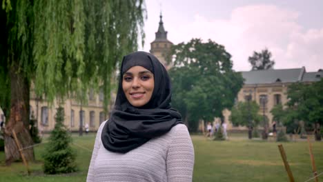 young sweet muslim girl in hijab is standing and smiling in daytime, in bad weather, watching at camera, building on background, religiuos concept