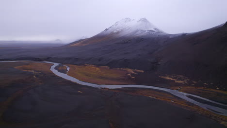 Fluss-überquert-Einen-Vulkanischen,-Schwarzen,-Schneebedeckten-Berg-In-Der-Wildnis-Islands