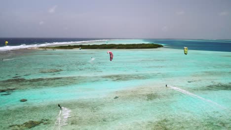 Kite-surfers-gliding-over-a-vibrant-tropical-coral-reef-in-clear-blue-waters,-aerial-view