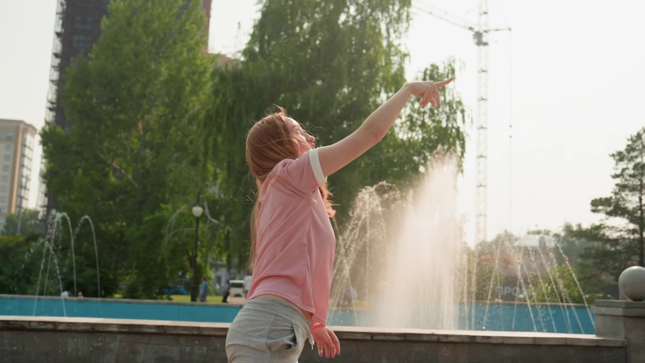 Young woman joyfully plays with paper airplane in outdoor park setting. Woman holds throws and watches plane as it flies away near water fountains wearing casual clothing