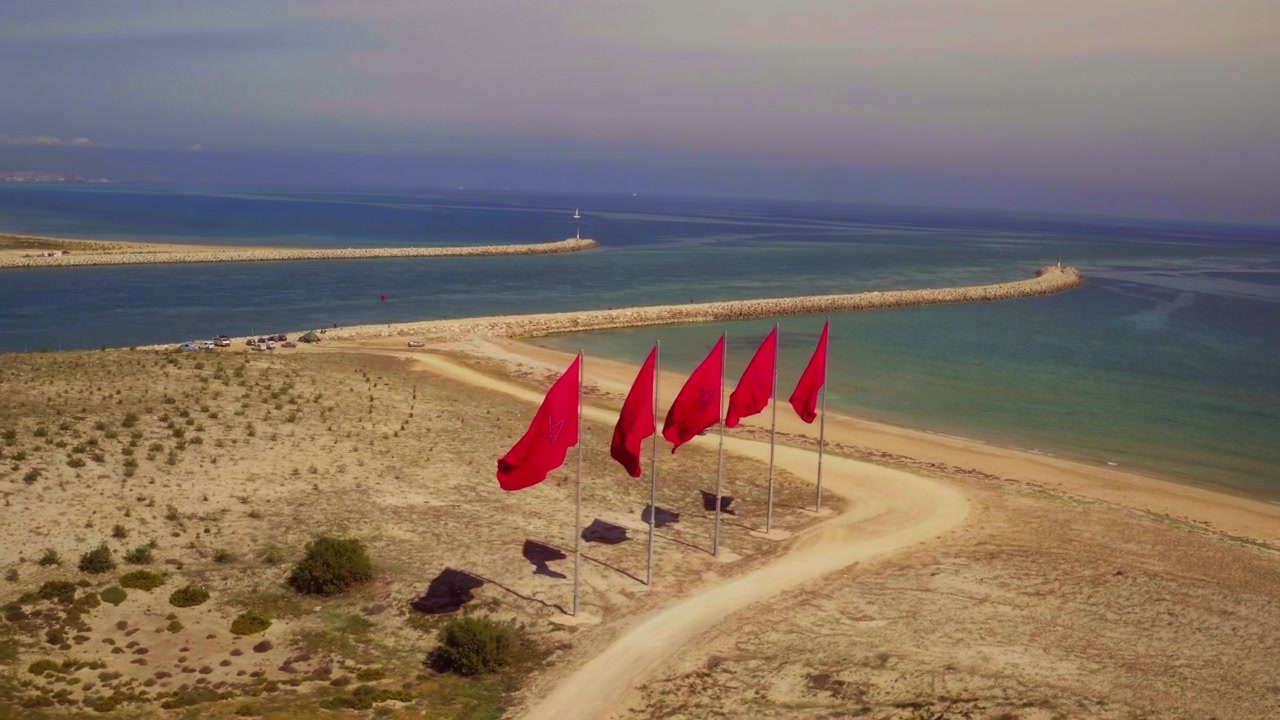Five red flags stake the sky above Morocco's golden sandy coastal spit.