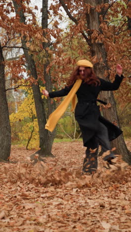 vista lateral de una dama en el bosque esparciendo hojas secas, con un largo abrigo negro, zapatos de lona y vaqueros negros, con un silenciador amarillo balanceándose