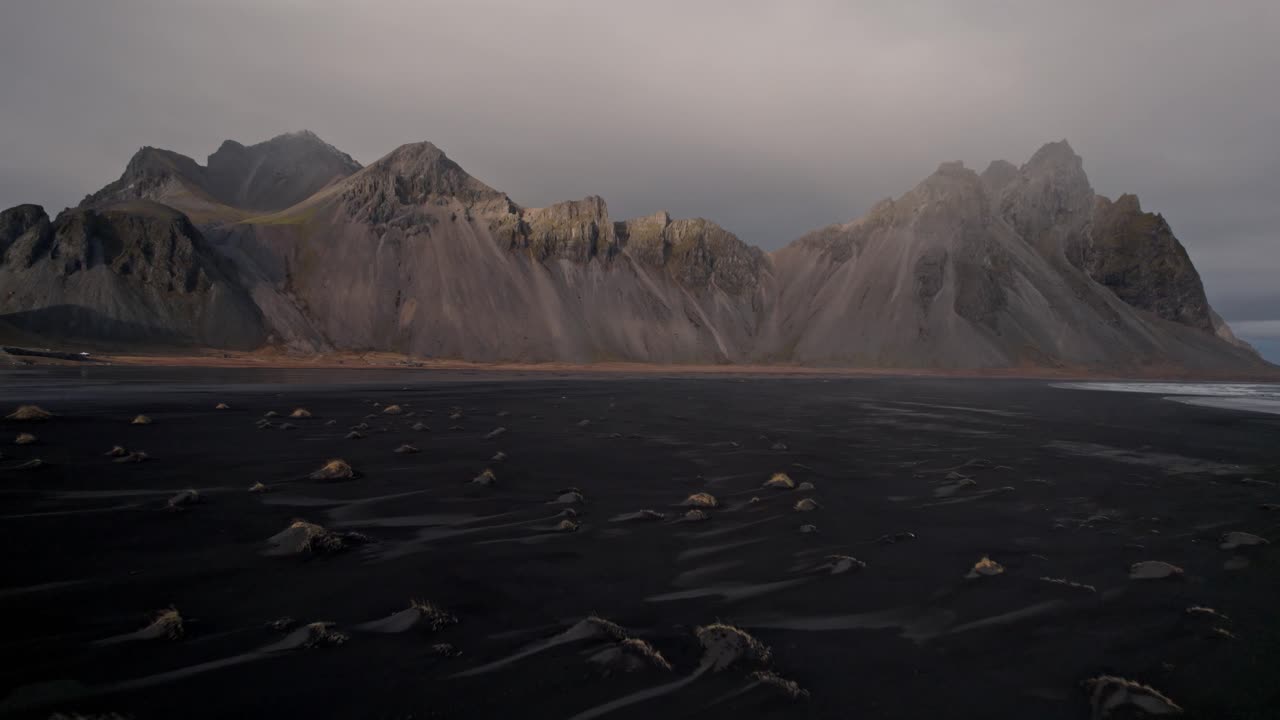 An evocative aerial shot captures the iconic Vestrahorn mountains rising dramatically behind the vast, volcanic black sand beach of Stokksnes on Iceland's southeastern coast.
