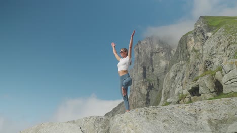 mujer en postura de árbol de yoga en un acantilado rocoso con cielo azul y montañas en lofoten, noruega, en cámara lenta