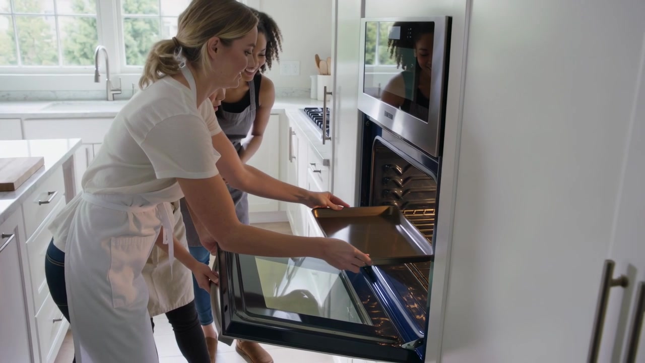 Lowering oven door, women in aprons sliding baking sheet into built-in oven in kitchen to bake