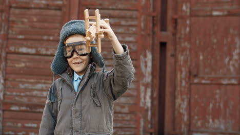 niño pelirrojo sonriente con sombrero y gafas jugando con un avión de juguete de madera y mirando la cámara