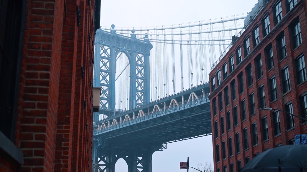 Nova Iorque, EUA, 25 de dezembro de 2025: Torre da Ponte de Manhattan emoldurada por paredes de armazéns de tijolos, DUMBO, Brooklyn. Vista de baixo ângulo da estrutura de suspensão de aço entre fachadas de tijolos vermelhos em um dia de neblina
