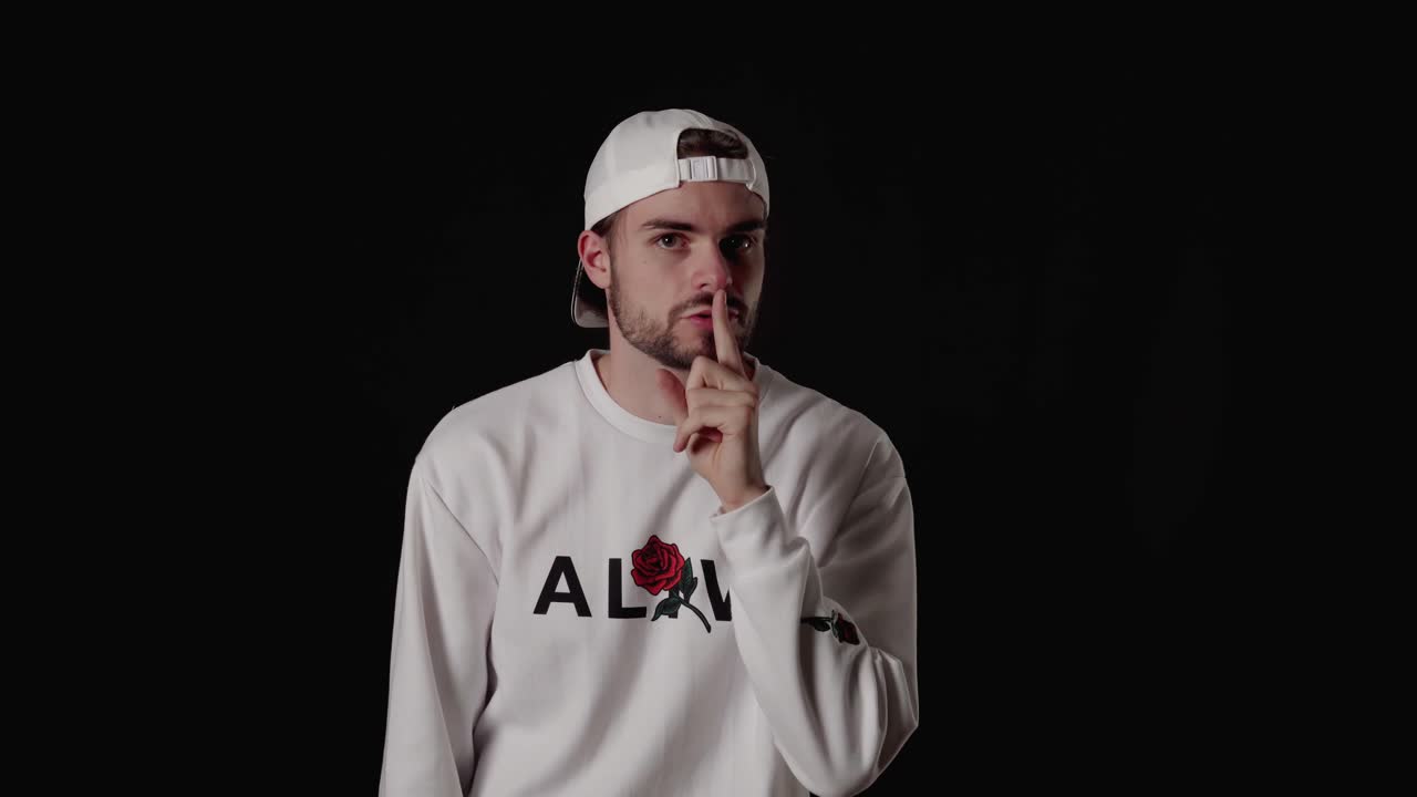 Trendy dvacetiletý muž utišuje do kamery, gesto ticha, široký záběr, černé pozadí.