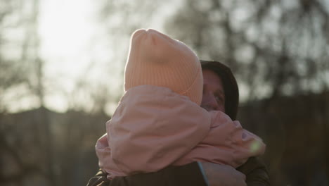 a little girl in a pink cap and jacket being carried by a man wearing a brown jacket, as she lovingly kisses him on the cheek