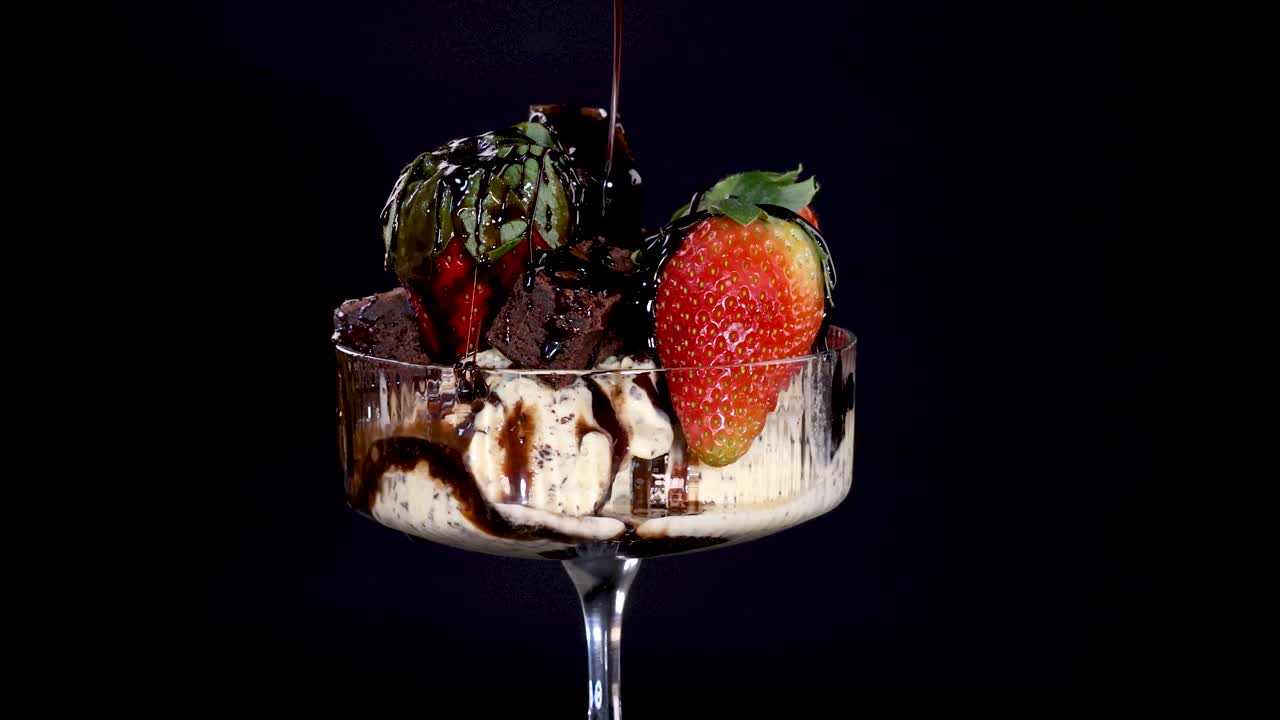 Rich chocolate sauce streams onto a glass dish of vanilla ice cream, fresh strawberries, chocolate chips, and brownie pieces against a dark background with dramatic lighting