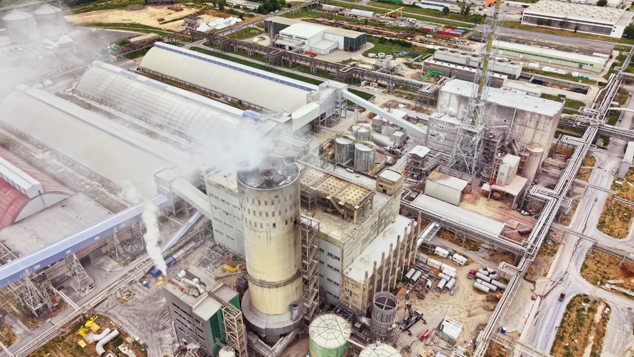 Power plant seen from above during the day with large buildings, pipes, and steam rising