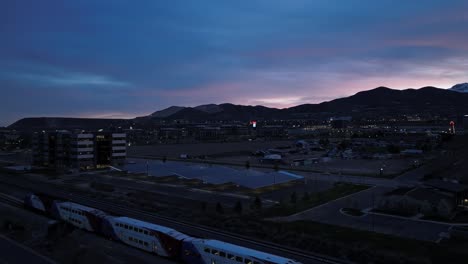 A-predawn-commuter-train-along-a-track-by-a-modern-office-building-with-a-solar-panel-parking-lot---aerial-flyover